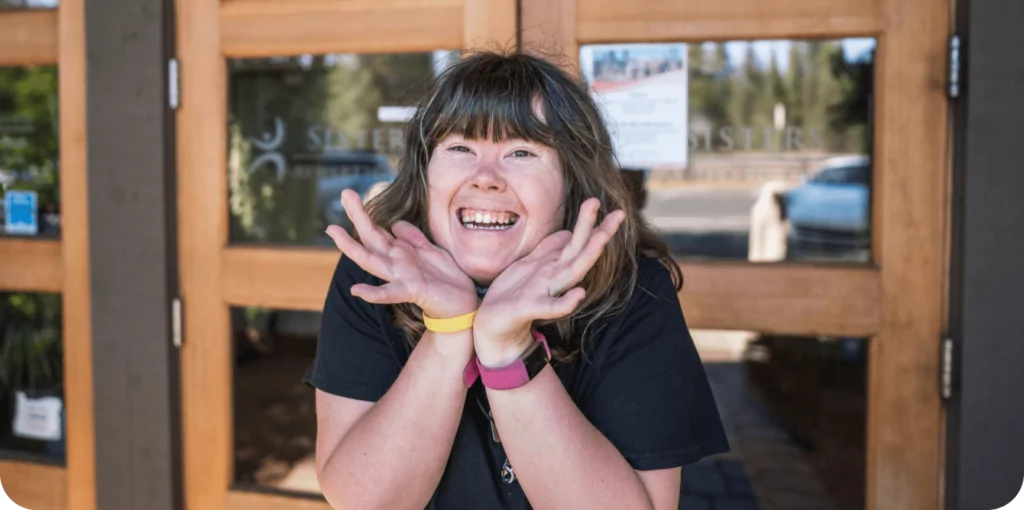 A developmentally disabled young woman sits at a table smiling at the camera. 