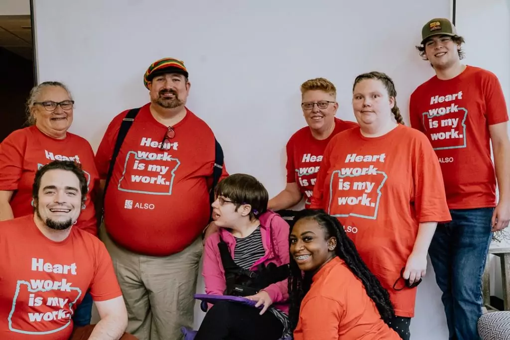 A group of ALSO direct support professionals stand together with developmentally disabled clients and smile at the camera. 