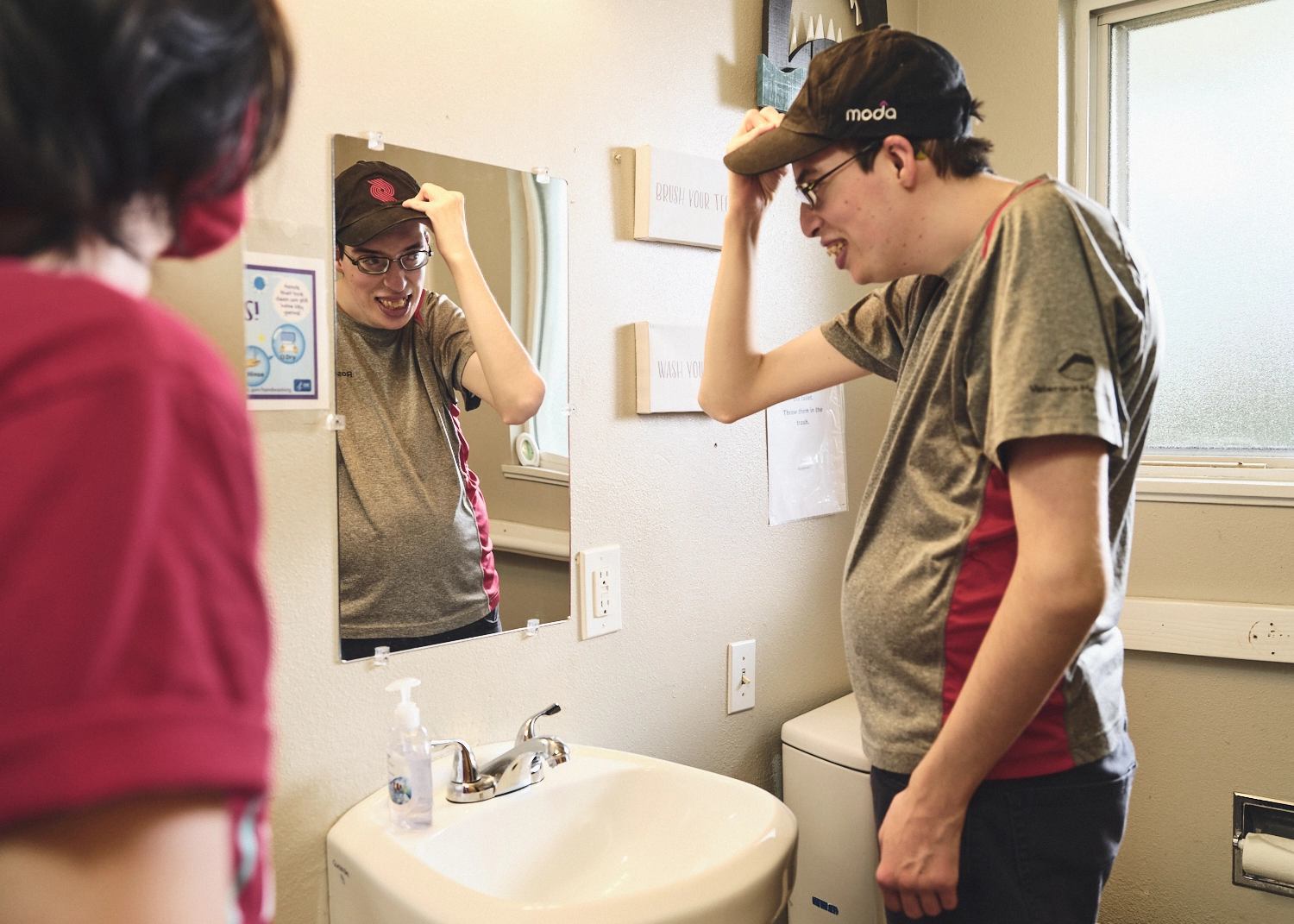 A developmentally disabled young man puts on a ball cap for work while looking in the mirror.