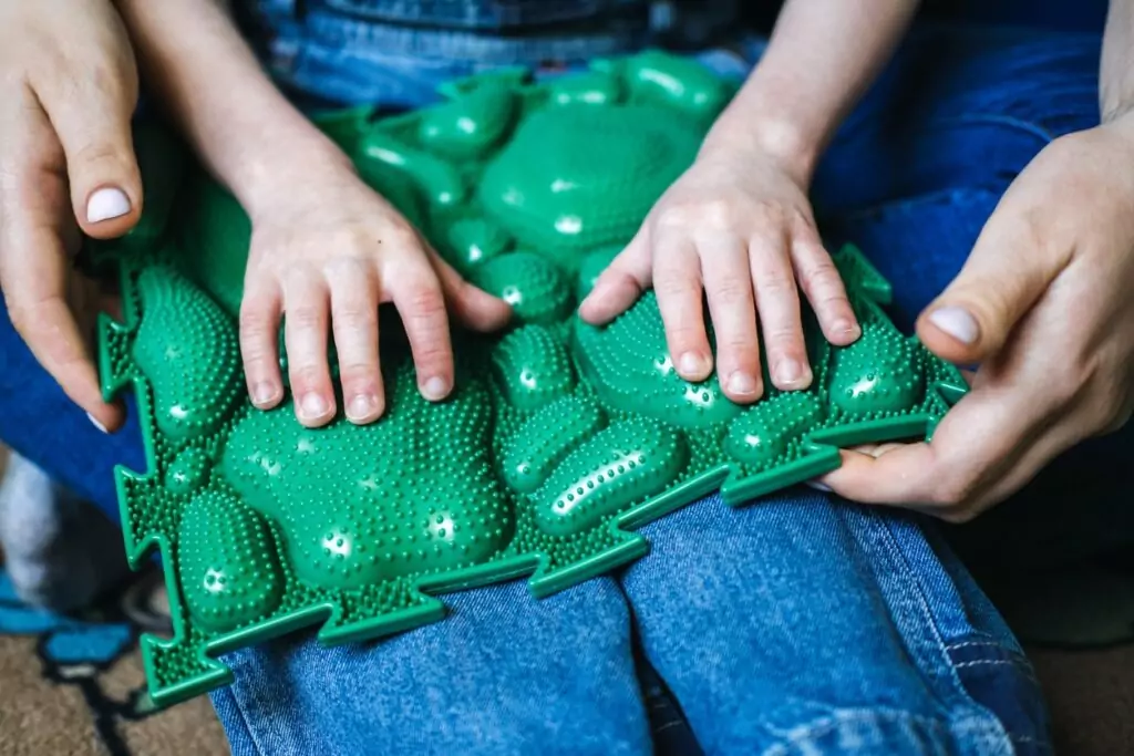 A boy sitting on lap of therapist engaging in sensory play with green textured plastic. 