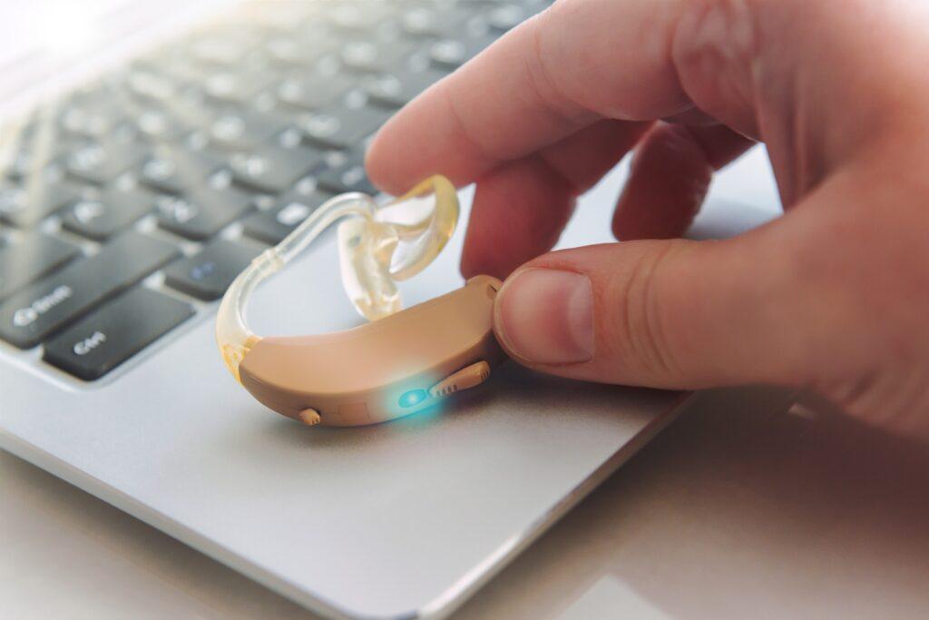Close up of a man's fingers placing a hearing aid on the edge of his laptop. 