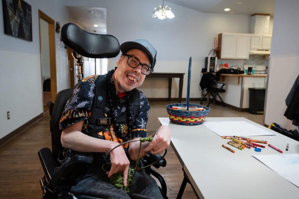 A young man with developmental disabilities in a wheelchair sits at a table and smiles at the camera.