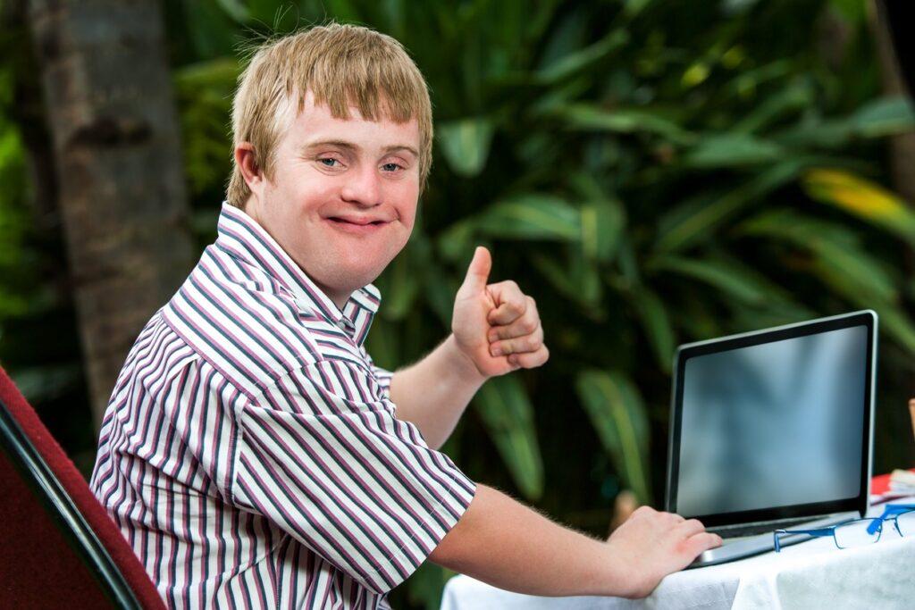 Developmentally disabled boy smiling and holding a thumbs up while sitting at a laptop. 