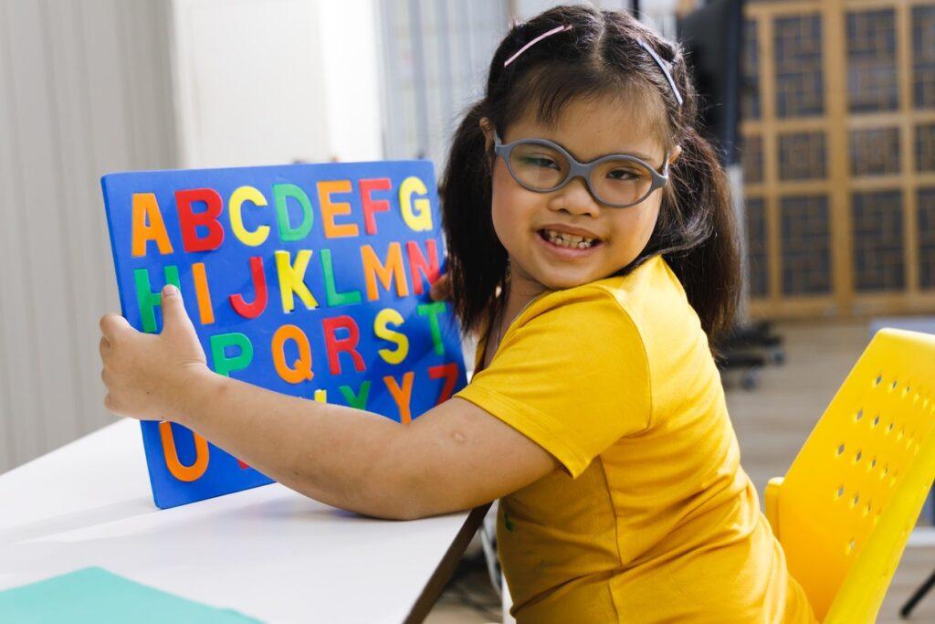A developmentally disabled girl in glasses holds up a chart of the alphabet. 