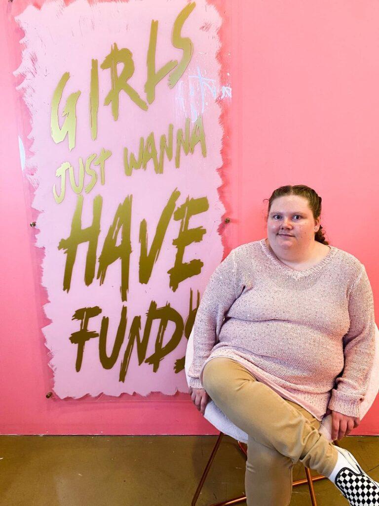 A developmentally disabled woman sits in front of a pink "Girls Just Wanna Haven Fund" sign at an event.