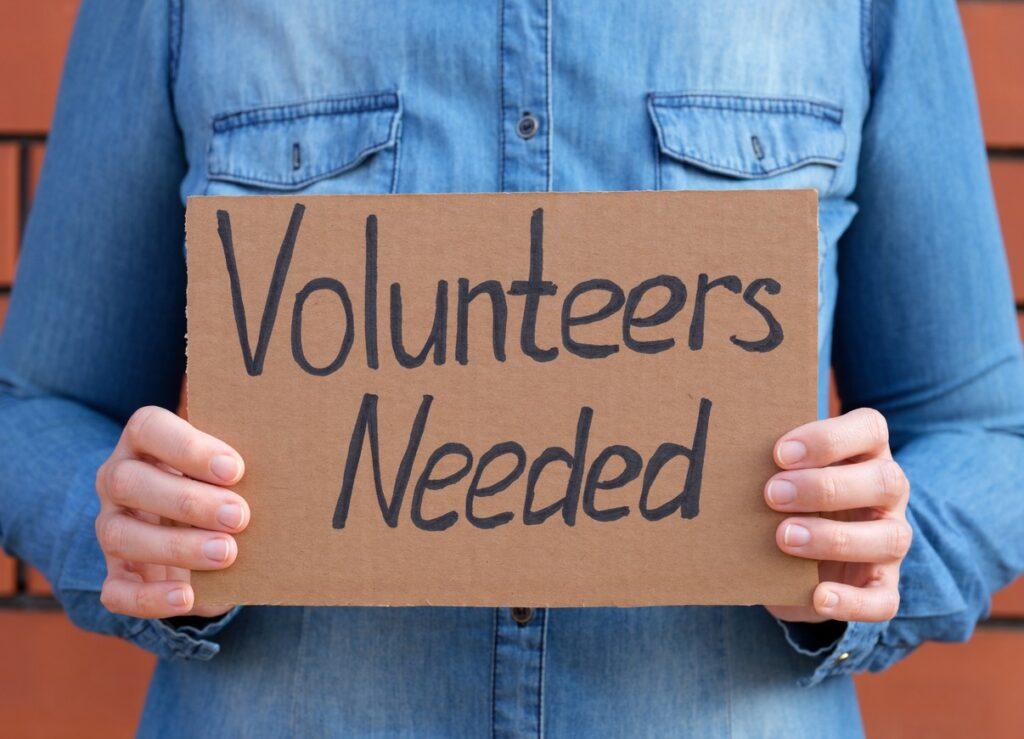 Close up of a person holding a handwritten "Volunteers Needed" sign.