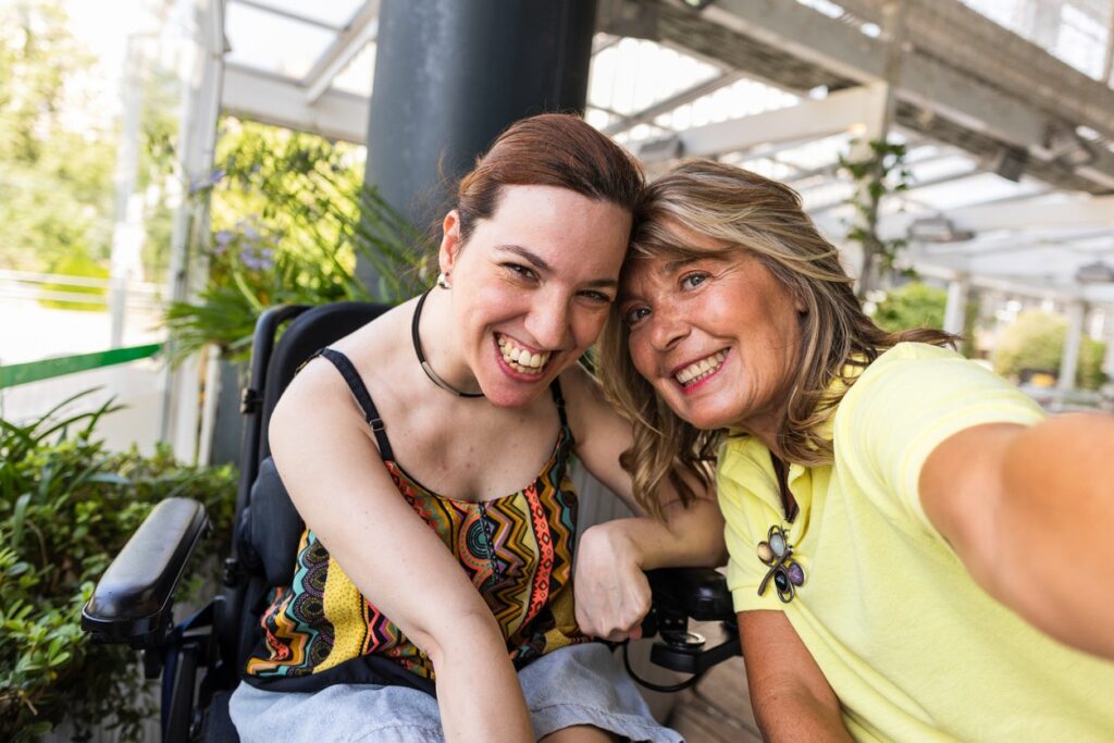 A mother and her adult disabled daughter, who is in a wheelchair, lean in together to take a selfie photo.