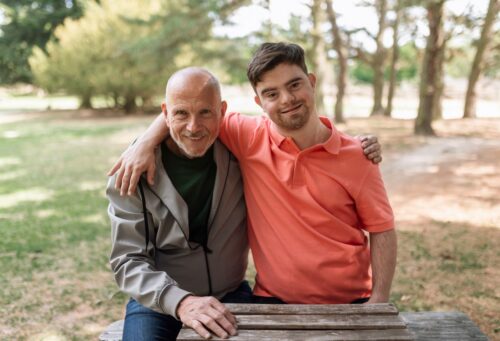 A senior father hugging his developmentally disabled son in a park and both are smiling at the camera.